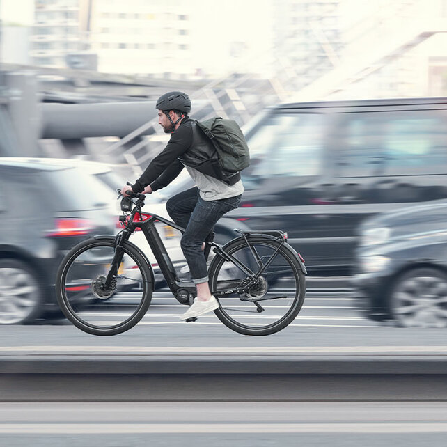 Un eBike piloto conduce por una carretera al lado Auto larga, todo excepto él está ligeramente borroso.