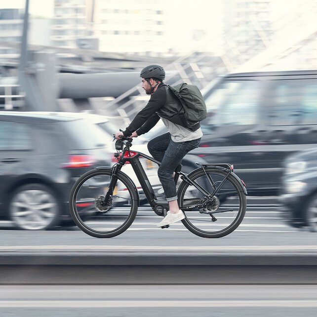 Ein eBike Fahrer fährt neben Autos eine Straße entlang, alles bis auf ihn ist leicht verschwommen.