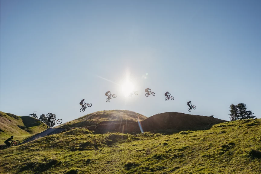 Shot from a low angle: a person jumps over a large dirt ramp beneath a blue sky and sun, with every phase of the jump visible. Grass is in the foreground.