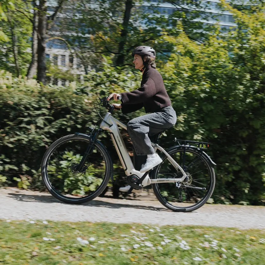 Žena jede na svém šedém eBike do kopce, míjí zelené živé ploty. Rozmazání pohybu naznačuje vysokou rychlost.