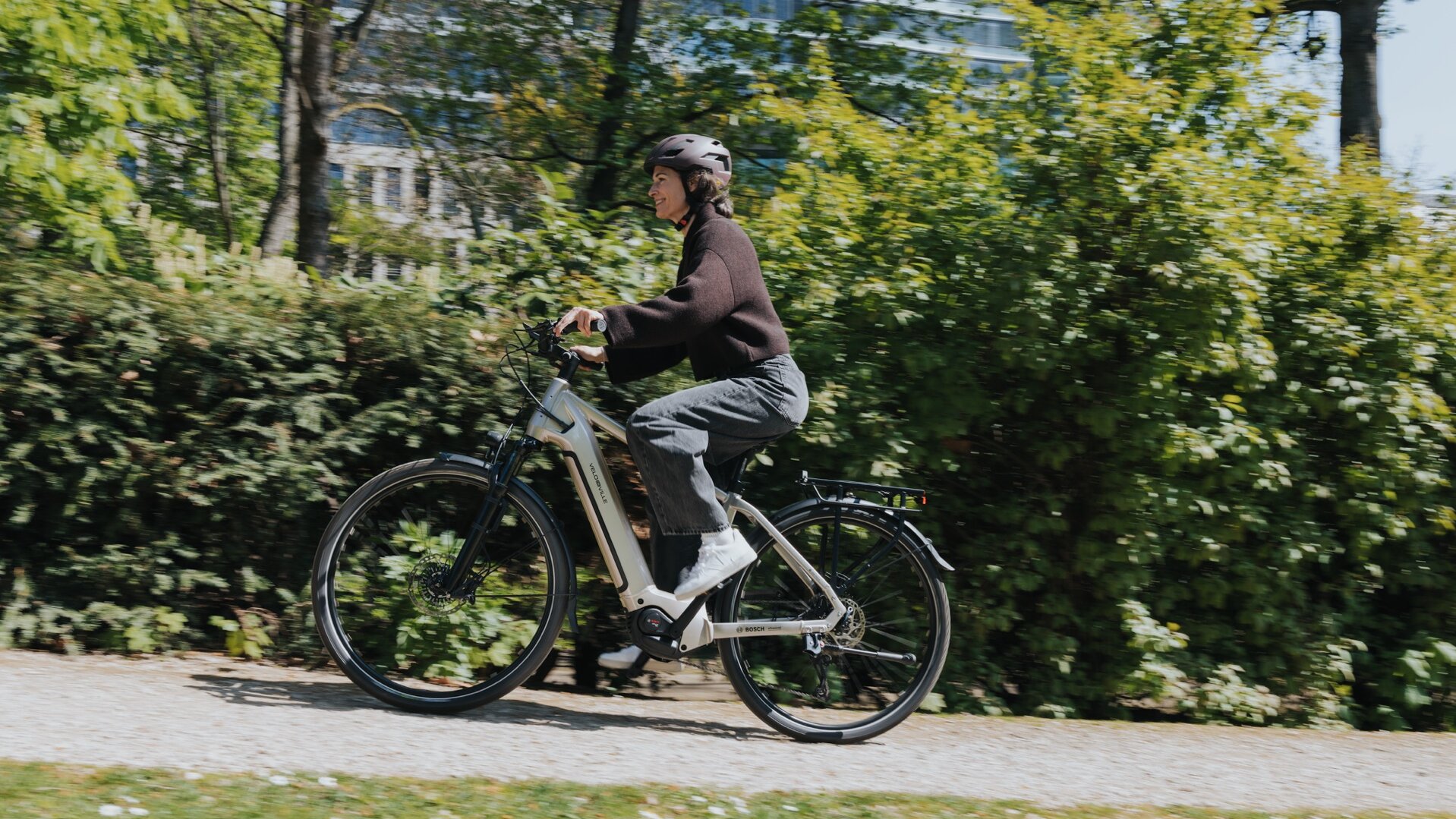 Žena jede na svém šedém eBike do kopce, míjí zelené živé ploty. Rozmazání pohybu naznačuje vysokou rychlost.