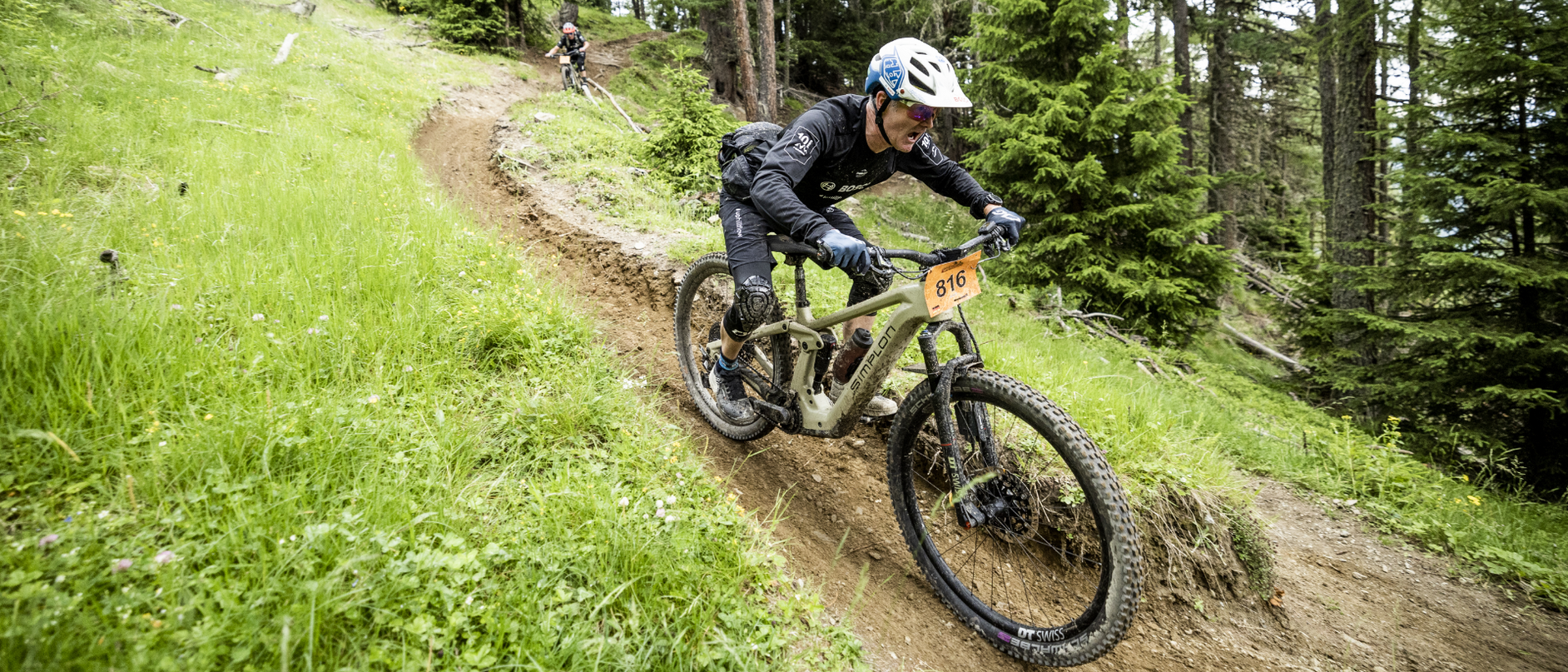 Ein Mann fährt auf einem eMountainbike einen erdigen Downhillpfad hinunter, der sich zwischen Wiese und Wald erstreckt. Im Hintergrund ist ein weiterer eMountainbikefahrer zu sehen. 