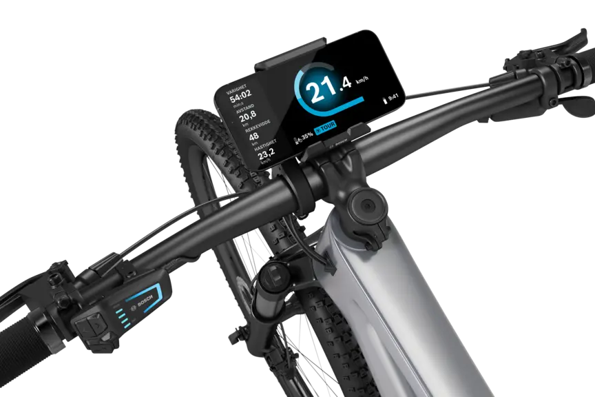 En sølvfarget elektrisk terrengsykkel med sort styre, isolert mot hvit bakgrunn. En SmartphoneGrip er montert på styret, og en smarttelefon er festet i den. Den åpne eBike Flow App viser fart i km/t, varighet i minutter, tilbakelagt distanse i kilometer, rekkevidde i kilometer og gjennomsnittshastighet. I tillegg vises batteristatus og kjøremodus.