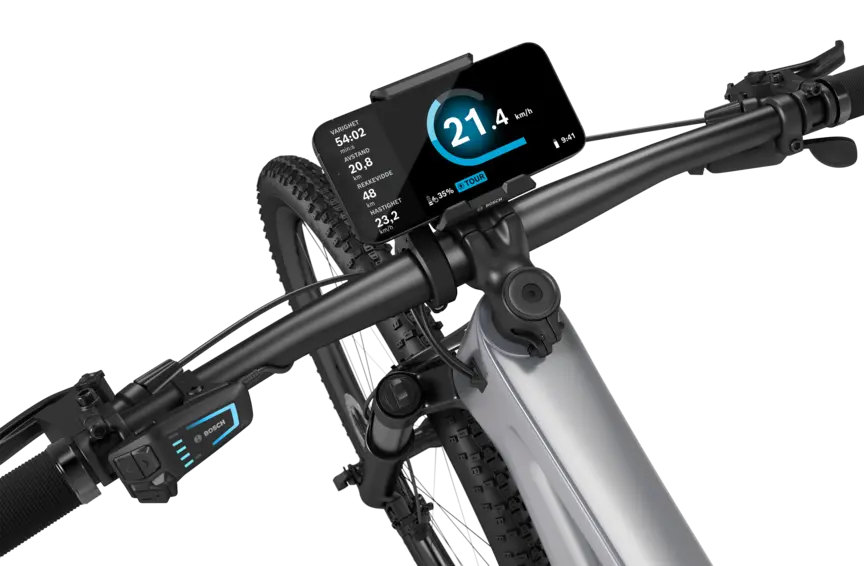 En sølvfarget elektrisk terrengsykkel med sort styre, isolert mot hvit bakgrunn. En SmartphoneGrip er montert på styret, og en smarttelefon er festet i den. Den åpne eBike Flow App viser fart i km/t, varighet i minutter, tilbakelagt distanse i kilometer, rekkevidde i kilometer og gjennomsnittshastighet. I tillegg vises batteristatus og kjøremodus.