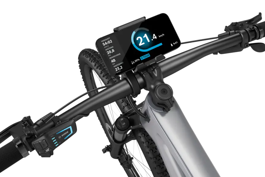 En sølvfarget elektrisk terrengsykkel med sort styre, isolert mot hvit bakgrunn. En SmartphoneGrip er montert på styret, og en smarttelefon er festet i den. Den åpne eBike Flow App viser fart i km/t, varighet i minutter, tilbakelagt distanse i kilometer, rekkevidde i kilometer og gjennomsnittshastighet. I tillegg vises batteristatus og kjøremodus.