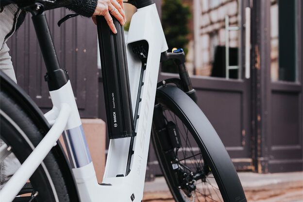 A person is removing the battery from a white e-bike. They are wearing a gray jacket and light-colored pants. In the background, you can see a building with large windows and a door.