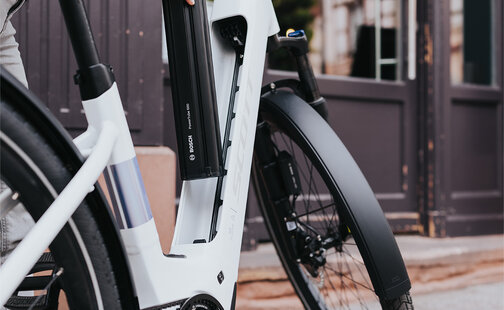 Una persona rimuove la batteria di una e-bike bianca. Indossa una giacca grigia e pantaloni chiari. Sullo sfondo si vede un edificio con grandi finestre e una porta.