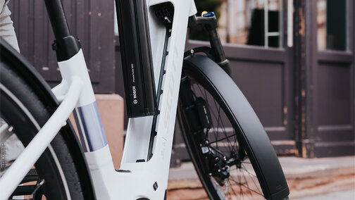 Eine Person entfernt den Akku eines weißen E-Bikes. Die Person trägt eine graue Jacke und helle Hosen. Im Hintergrund sind ein Gebäude mit großen Fenstern und eine Tür zu sehen.