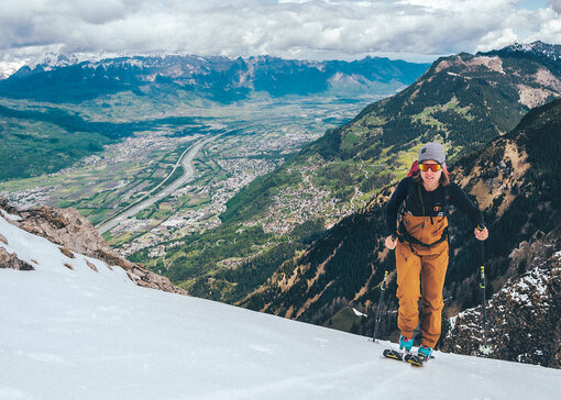 Une femme pratique le ski de randonnée sur une montagne enneigée. En arrière-plan, une ville s’étend en contrebas.