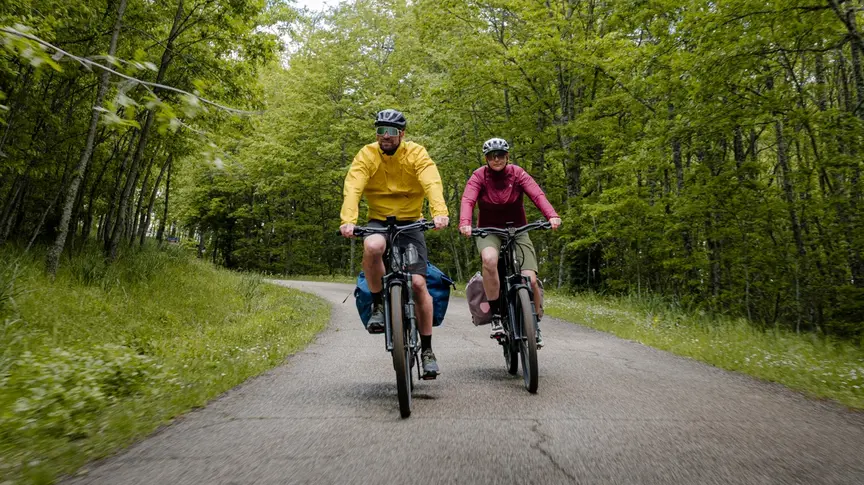 Ein Mann in einer gelben und eine Frau in einer roten Jacke fahren auf einer Straße mit ihren bepackten Trekking eBikes durch einen üppigen Laubwald.