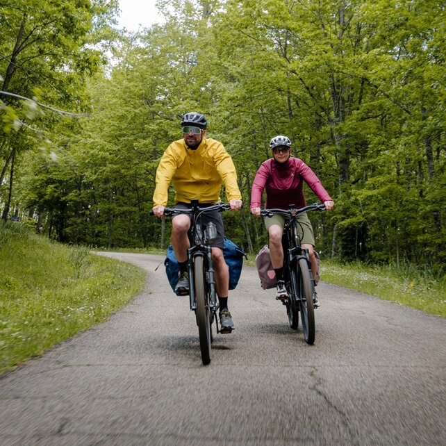 A man in a yellow jacket and a woman in a red jacket ride their packed trekking eBikes along a road through a lush deciduous forest.