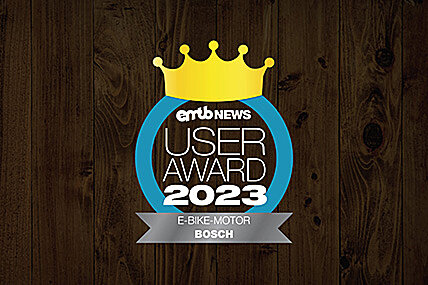 Icon with the text "emtb NEWS USER AWARD 2023": A golden crown above the text, enclosed by a blue circle. "E-BIKE-MOTOR BOSCH" is written at the bottom. The background has a dark wood finish.