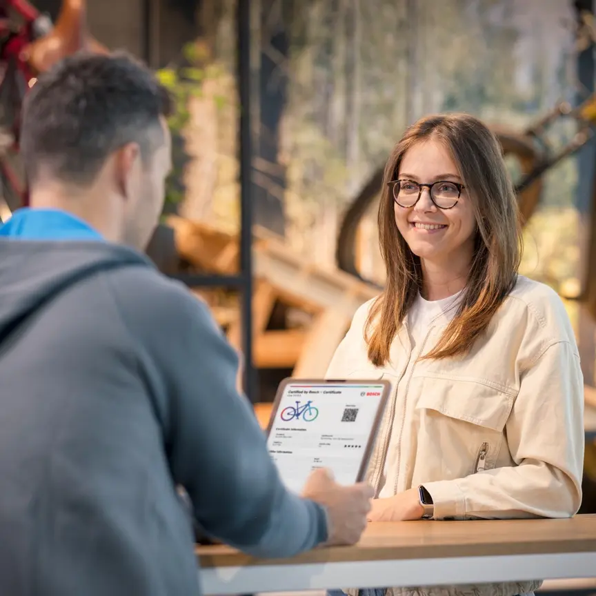 Ein Mann zeigt einer lächelnden Mitarbeiterin in einem Fahrradgeschäft ein "Certified by Bosch"-Zertifikat auf einem Tablet.