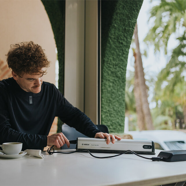 A mountain biker sits at a table on a café terrace and connects his Bosch eBike battery, the PowerTube 800, to the 12A charger. Next to the battery is a coffee cup, with palm trees and Mediterranean architecture visible in the background.