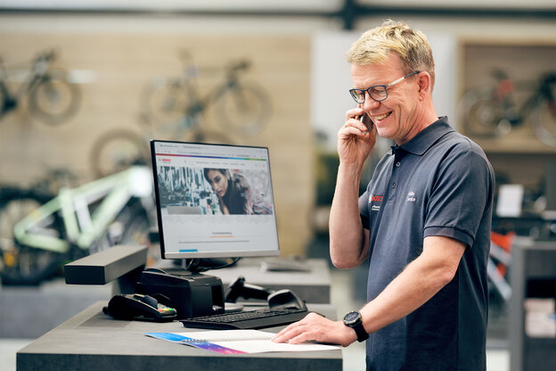 eBike-experten står vid sitt skrivbord i cykelaffären. På datorn är Bosch-webbplatsen öppen medan han pratar i telefon med Boschs kundtjänst.