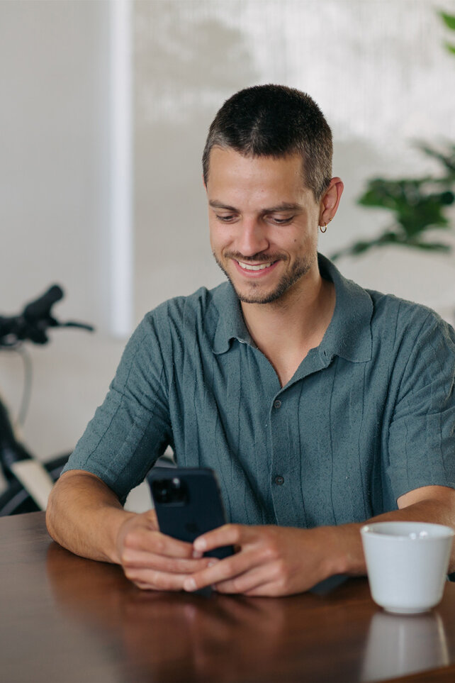 A man is sitting at a table, smiling as he looks at his smartphone.