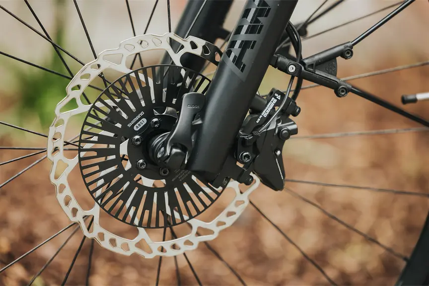 Close-up of a bicycle disc brake. The brake is mounted on a black bicycle fork, and the background is out of focus with brown and green tones.