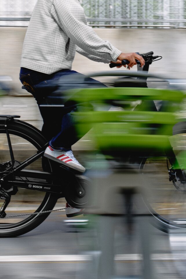 foto de una ciclista en un eBike negro con un motor Bosch en el cubo. La conductora está enfocada mientras pasa junto a una serie de objetos verdes brillantes que se difuminan en franjas dinámicas debido al rápido movimiento, transmitiendo una fuerte sensación de velocidad.