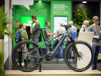 Photo of a blue eMountain bike on display at a trade fair, with booths visible in the background.