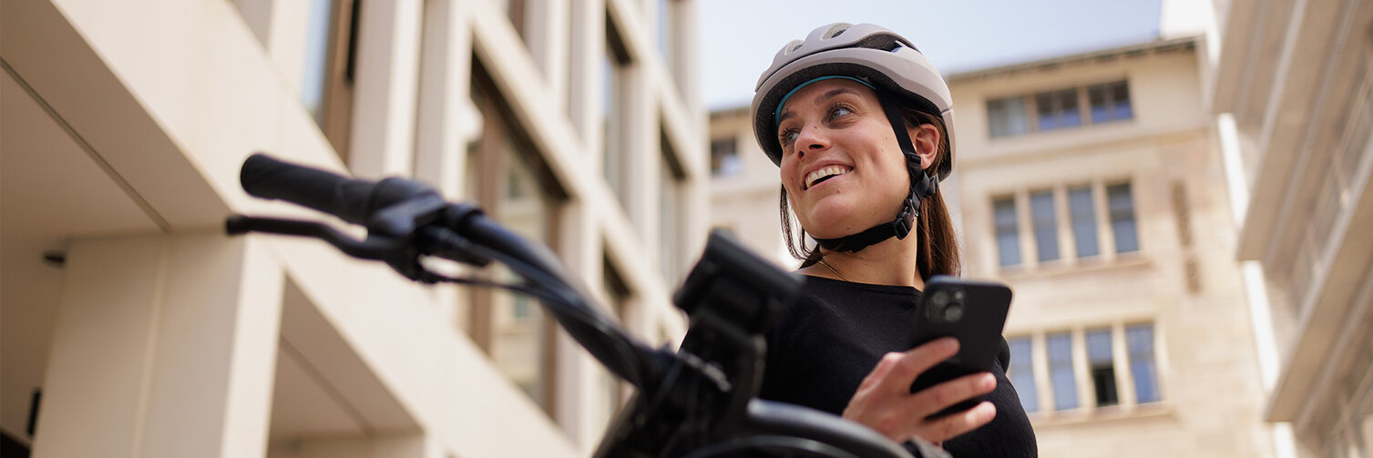 A woman wearing a bicycle helmet looks to the side and smiles while holding her smartphone. In the background, buildings are visible, and the bicycle handlebars are blurred in the foreground.