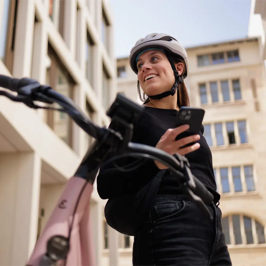 Frau mit Fahrradhelm schaut zur Seite und lächelt, während sie ihr Smartphone in der Hand hält. Im Hintergrund sind Gebäude zu sehen, und im Vordergrund unscharf der Fahrradlenker.