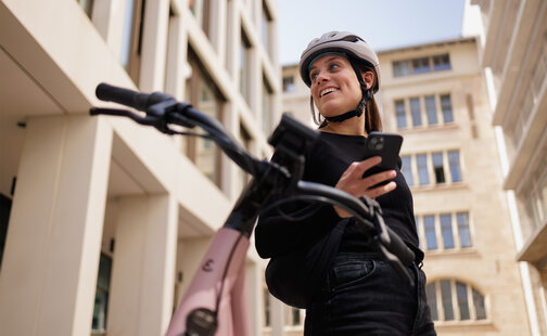 Frau mit Fahrradhelm schaut zur Seite und lächelt, während sie ihr Smartphone in der Hand hält. Im Hintergrund sind Gebäude zu sehen, und im Vordergrund unscharf der Fahrradlenker.
