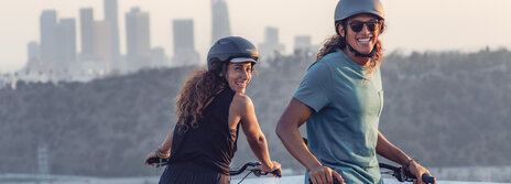 A man and a woman take a break with their bicycles at a scenic spot. The man leans back relaxed against the frame, while the woman looks over her shoulder. Both of them are smiling.