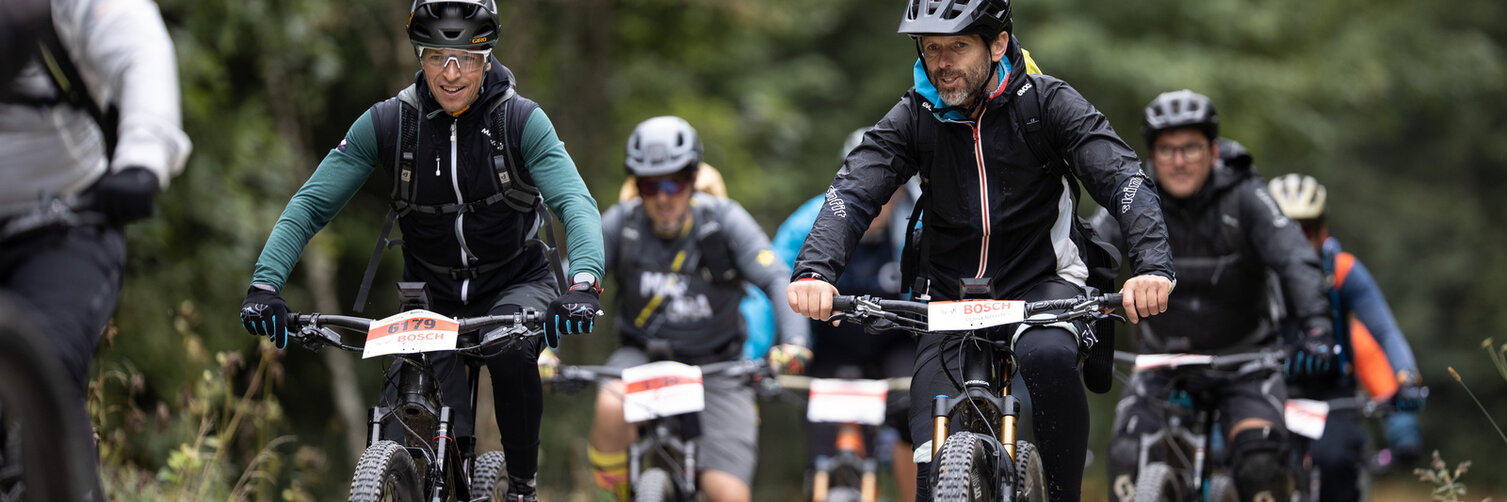 Eine Gruppe Radfahrer in Ausrüstung mit Startnummern an den Lenkern ist zu sehen. Im Hintergrund sind Bäume.