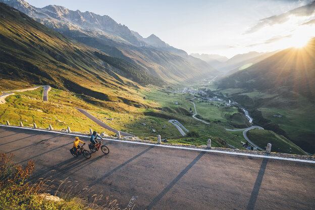 Deux conducteurs de VAE roulent le long d’une route au lever du soleil, avec une vallée et un village en contrebas, et des montagnes à l’arrière-plan.