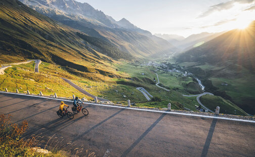 Due conducenti di eBike percorrono una strada all'alba; sotto di loro si apre una valle con un villaggio e sullo sfondo si vedono delle montagne.