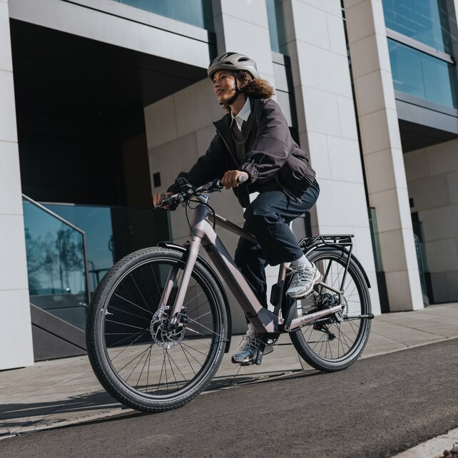 An eBiker wearing a helmet rides along an asphalt path beside the façade of a large, modern building.