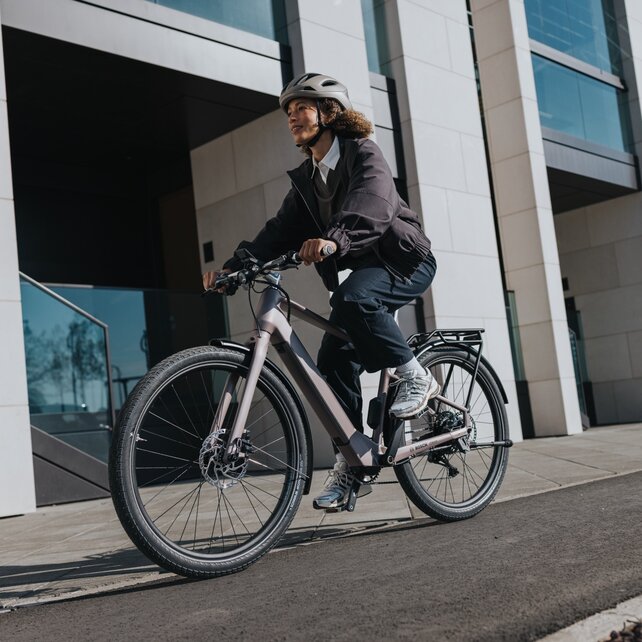 An eBiker wearing a helmet rides along an asphalt path beside the façade of a large, modern building.