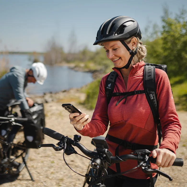 A woman wearing a red jacket and helmet sits on her e-bike, smiling as she looks at her smartphone. In the background, a man by the lake is adjusting his luggage bag.