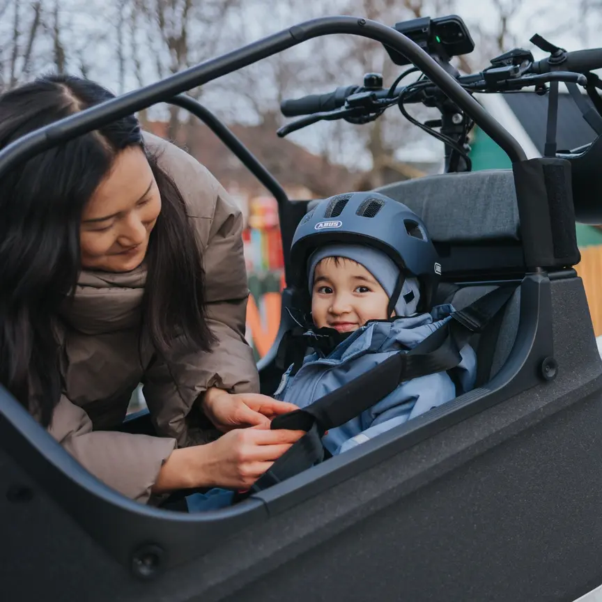 Eine Person sichert ein Kind in einem Kindersitz auf einem Lasten-E-Bike. Das Kind trägt einen Helm und warme Kleidung. Im Hintergrund sind Häuser und Bäume zu sehen.