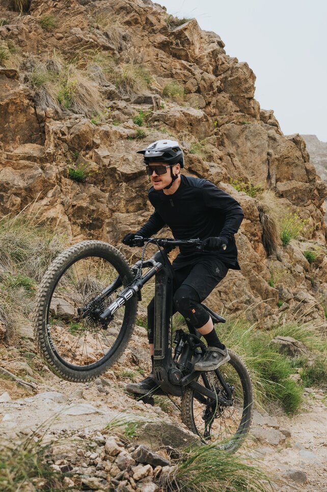 Un eMountainbiker affronta una salita ripida su un sentiero stretto che attraversa un paesaggio accidentato e roccioso. Sullo sfondo, un ampio canyon asciutto si estende sotto un cielo luminoso.
