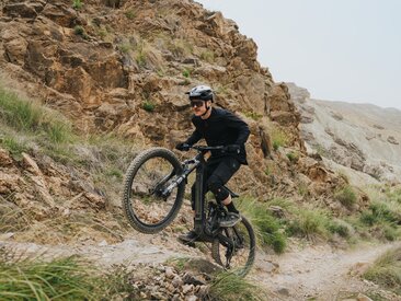 An eMountain biker rides steeply upward on a narrow path through a rugged, rocky landscape. In the background, a vast, dry canyon stretches out beneath a bright sky.