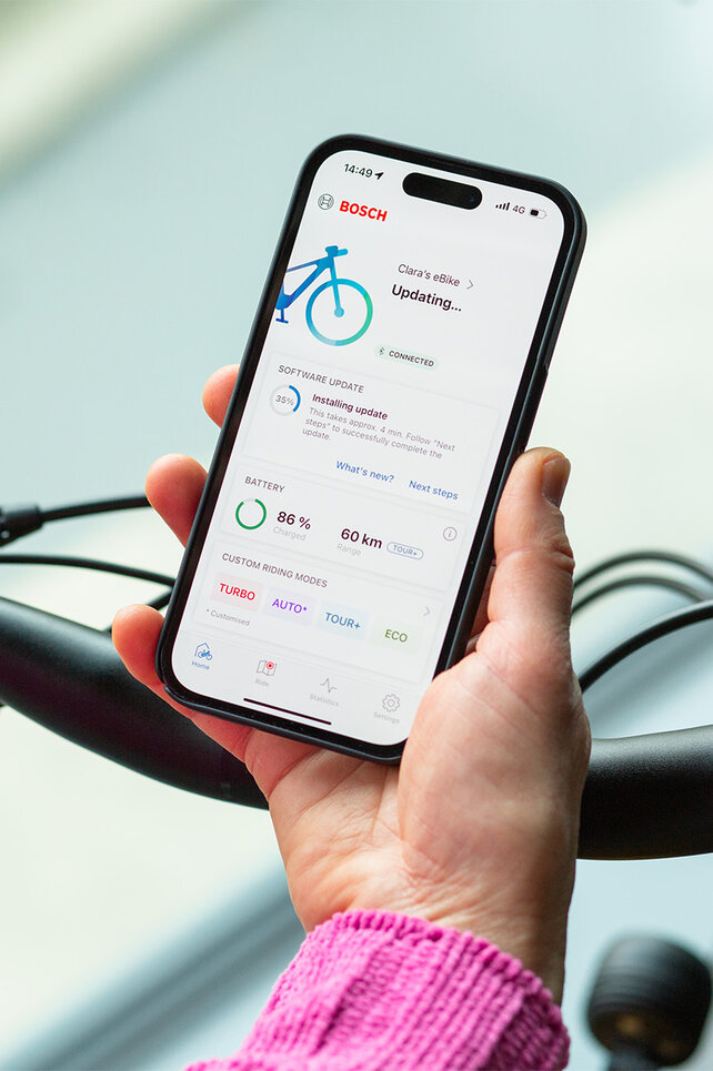 A person holds a smartphone near their eBike and installs updates using the smartphone app.