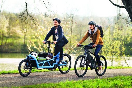 Two men are cycling their pedelecs through a park, one on a light blue cargo eBike, the other on a black eMountain bike