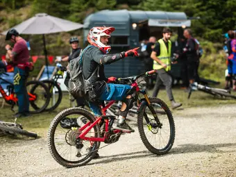 A mountain biker on an eMountain bike is seen from a diagonal angle from behind, pointing to the right with an outstretched arm and index finger. In the background, additional people and bicycles are visible.