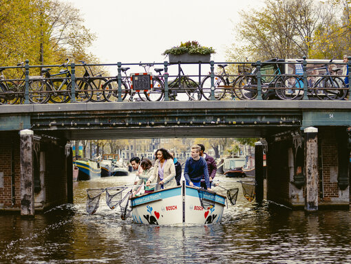 Meerdere mensen zitten op een kleine boot op een kanaal en vissen met schepnetten dingen uit het water terwijl ze onder een brug door varen.