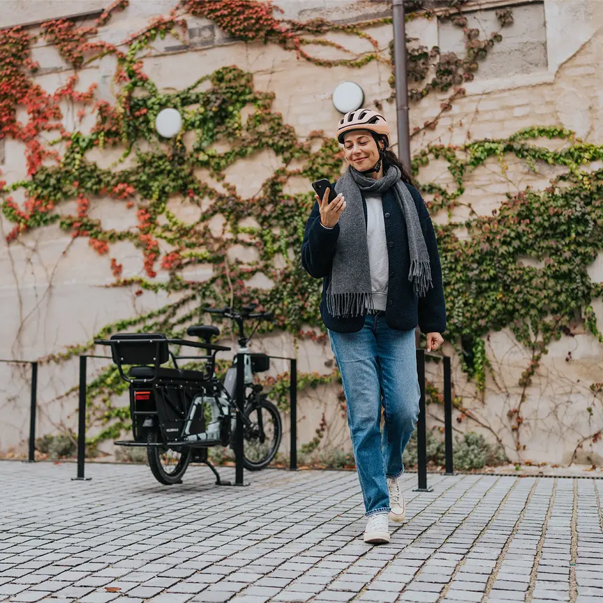 Eine Frau mittleren Alters hat gerade ihr eBike zum Parken abgestellt und entfernt sich vom eBike. In der Hand hält sie ein Smartphone.