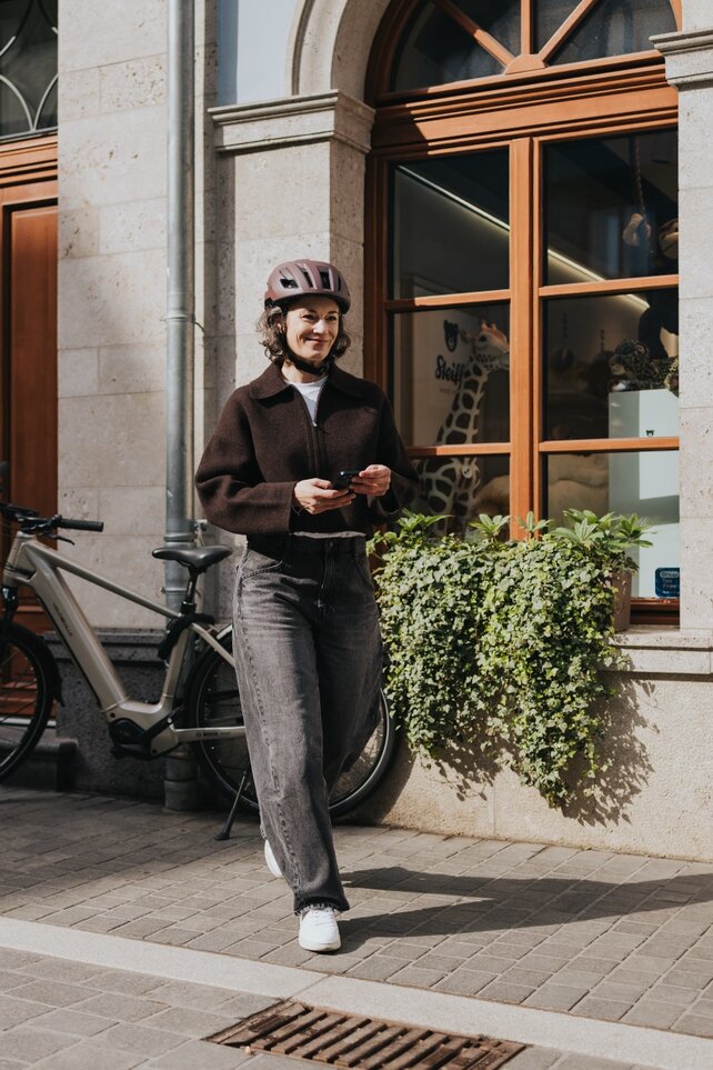 Una donna con casco da bicicletta cammina sorridente su un marciapiede in città, tenendo lo smartphone in mano. Il tuo eBike moderno è parcheggiato accanto a una vetrina, valorizzando uno stile di vita urbano e attivo.