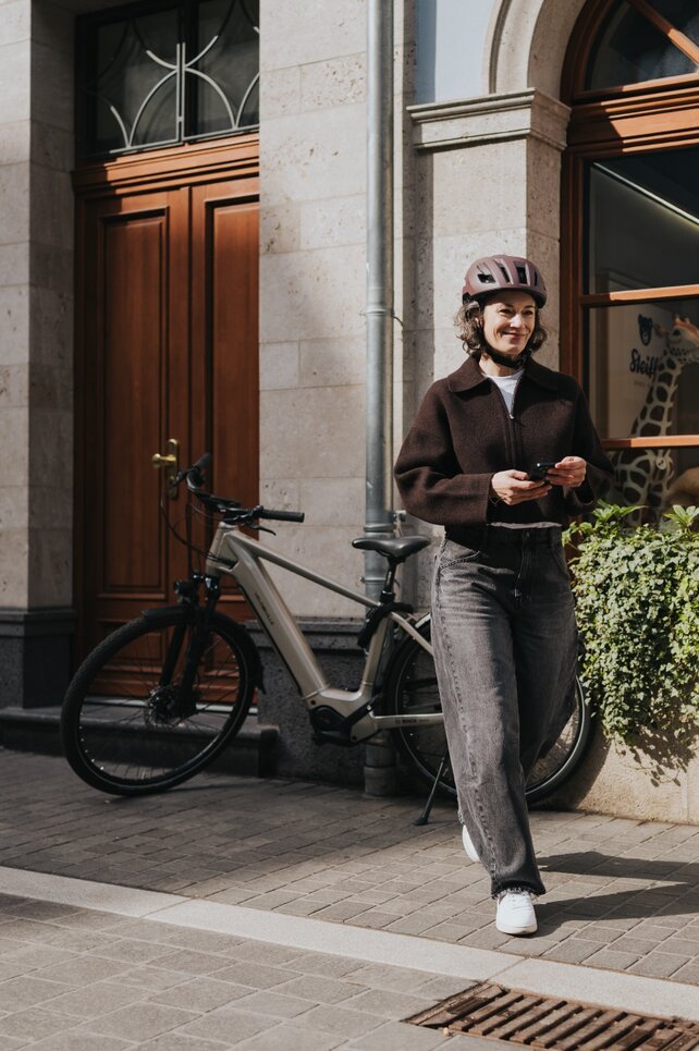Eine Frau mit Fahrradhelm geht lächelnd auf einem Bürgersteig in der Stadt und hält ihr Smartphone in der Hand. Ihr modernes eBike ist neben einem Schaufenster geparkt, was einen urbanen und aktiven Lebensstil unterstreicht.     