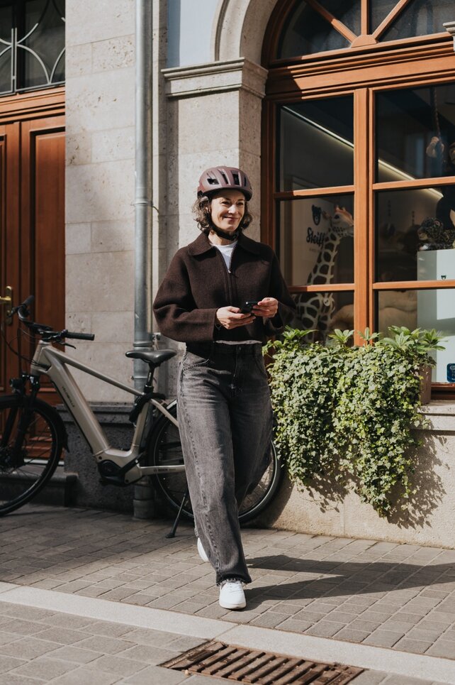 A woman wearing a bicycle helmet walks through the city, smiling and holding her smartphone. Her modern eBike is parked by a shop window, emphasizing an urban and active lifestyle.