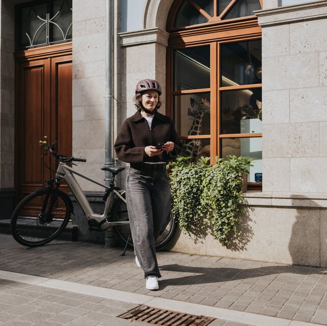 Une femme portant un casque de vélo marche sur un trottoir en ville, tout en affichant un sourire et tenant son smartphone. Votre VAE moderne est garé près d’une vitrine, soulignant un mode de vie urbain et dynamique.
