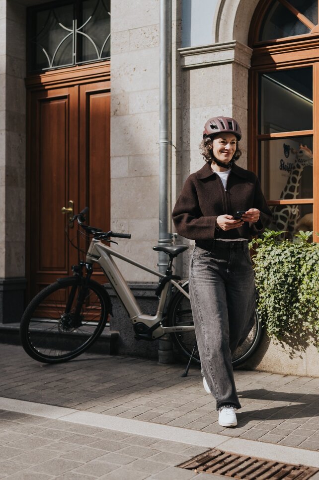 En kvinna med cykelhjälm går leende på en trottoar i staden och håller sin smartphone i handen. Din moderna elcykel står parkerad bredvid ett skyltfönster och framhäver en urban och aktiv livsstil.
