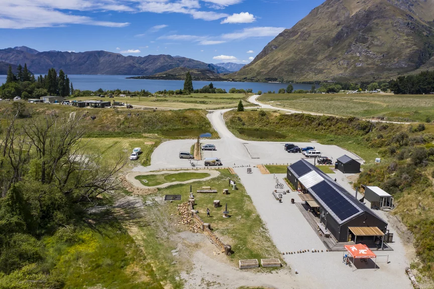An aerial view of the terrain shows a large building, a parking lot with cars, and, in the distance, a lake with mountains.