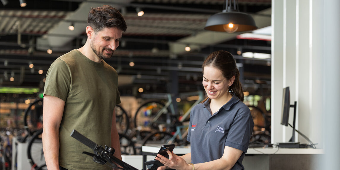 Two people are standing in a bicycle shop. One person is wearing an olive green T-shirt and black pants, while the other is dressed in a gray polo shirt with the Bosch logo and holding a smartphone. In the background, you can see several bicycles and modern furnishings.