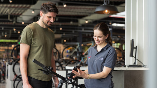 Two people are standing in a bicycle shop. One person is wearing an olive green T-shirt and black pants, while the other is dressed in a gray polo shirt with the Bosch logo and holding a smartphone. In the background, you can see several bicycles and modern furnishings.