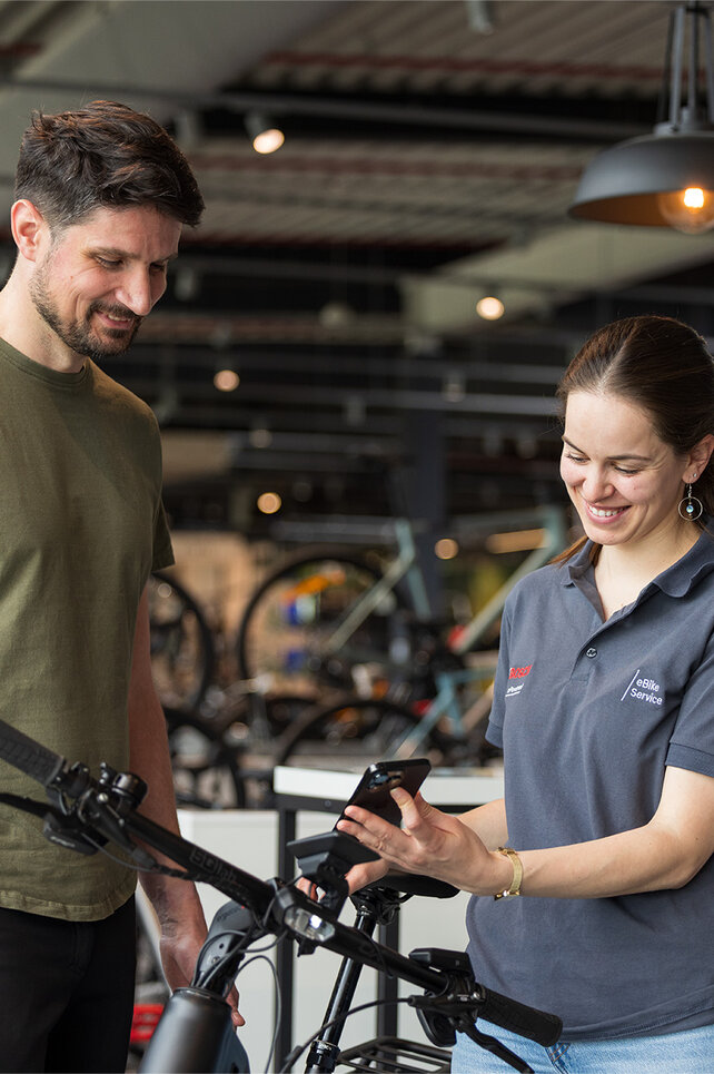 Zwei Personen stehen in einem Fahrradgeschäft. Eine Person trägt ein olivgrünes T-Shirt und schwarze Hosen, die andere Person trägt ein graues Poloshirt mit Bosch-Logo und hält ein Smartphone. Im Hintergrund sind mehrere Fahrräder und eine moderne Einrichtung zu sehen.